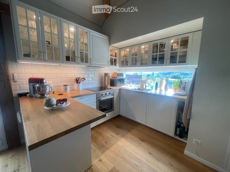 Modern kitchen with white cabinets, wooden countertop, and stainless steel appliances. Features an island with a mixer, bowl, and fruit, and a window overlooking a garden.