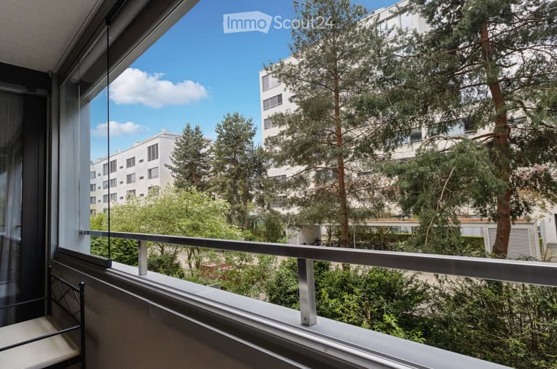 Balcony with metal railing, overlooking a tree-lined urban area with buildings and greenery.