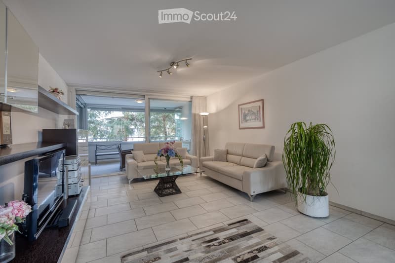 Modern living room with two beige sofas, a glass coffee table, a potted plant, and a TV on a stand. Large windows offer a view of the balcony and outdoor area.