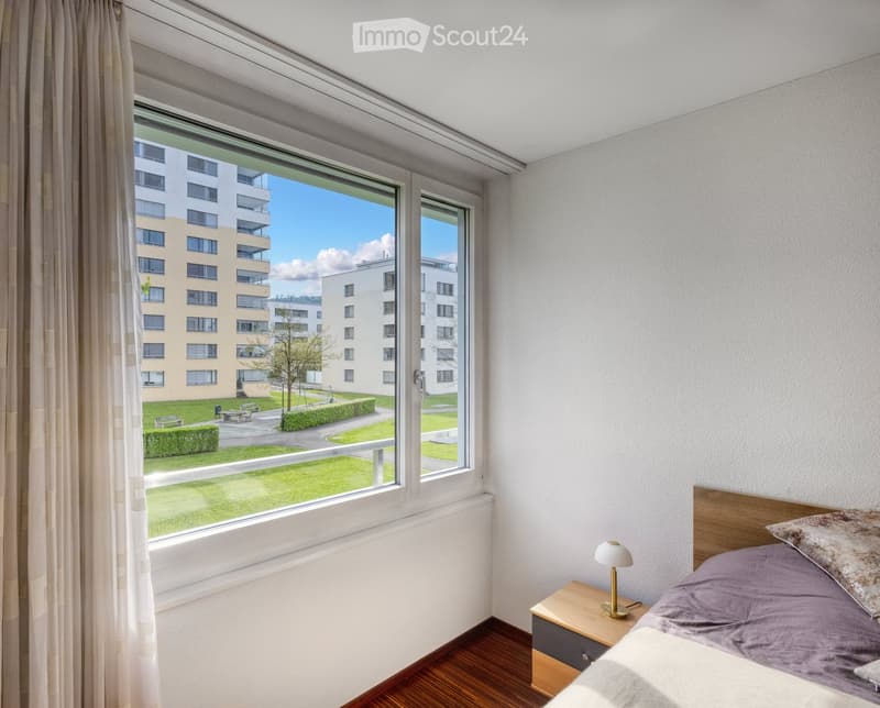 Bright bedroom with wooden flooring, large window offering a view of a green park and modern buildings, featuring a bed with pillows, a nightstand, and a lamp.