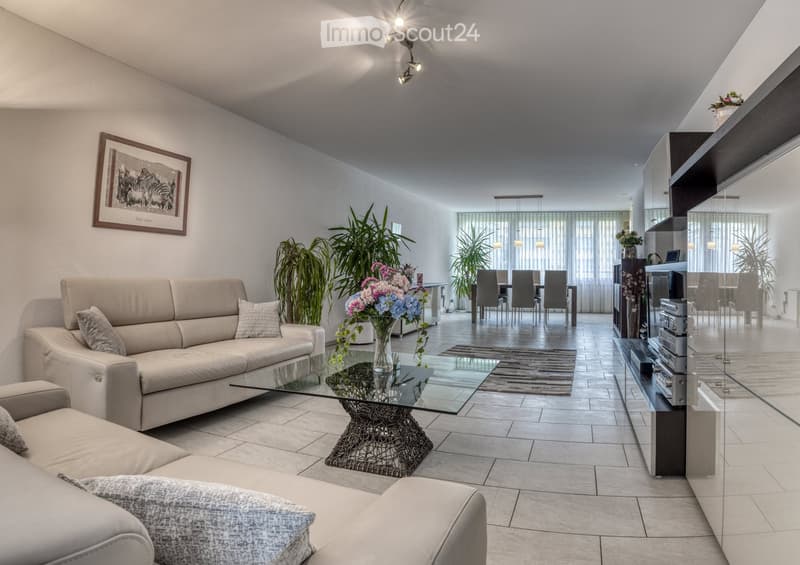 Modern living room with a beige sofa, glass coffee table, dining area with chairs and table, and decorative plants.
