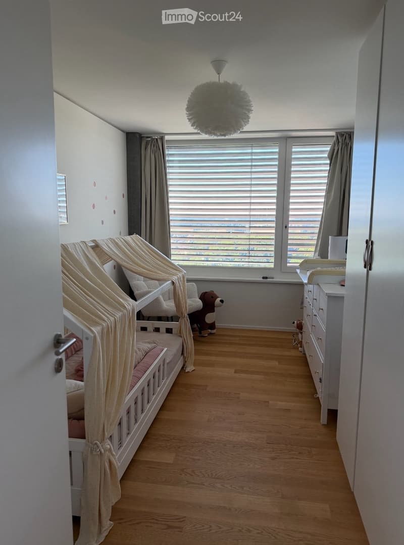 Bright, modern children's bedroom with a wooden floor, large window with blinds, a white bed with a beige canopy, a dresser, and a plush toy on the floor.