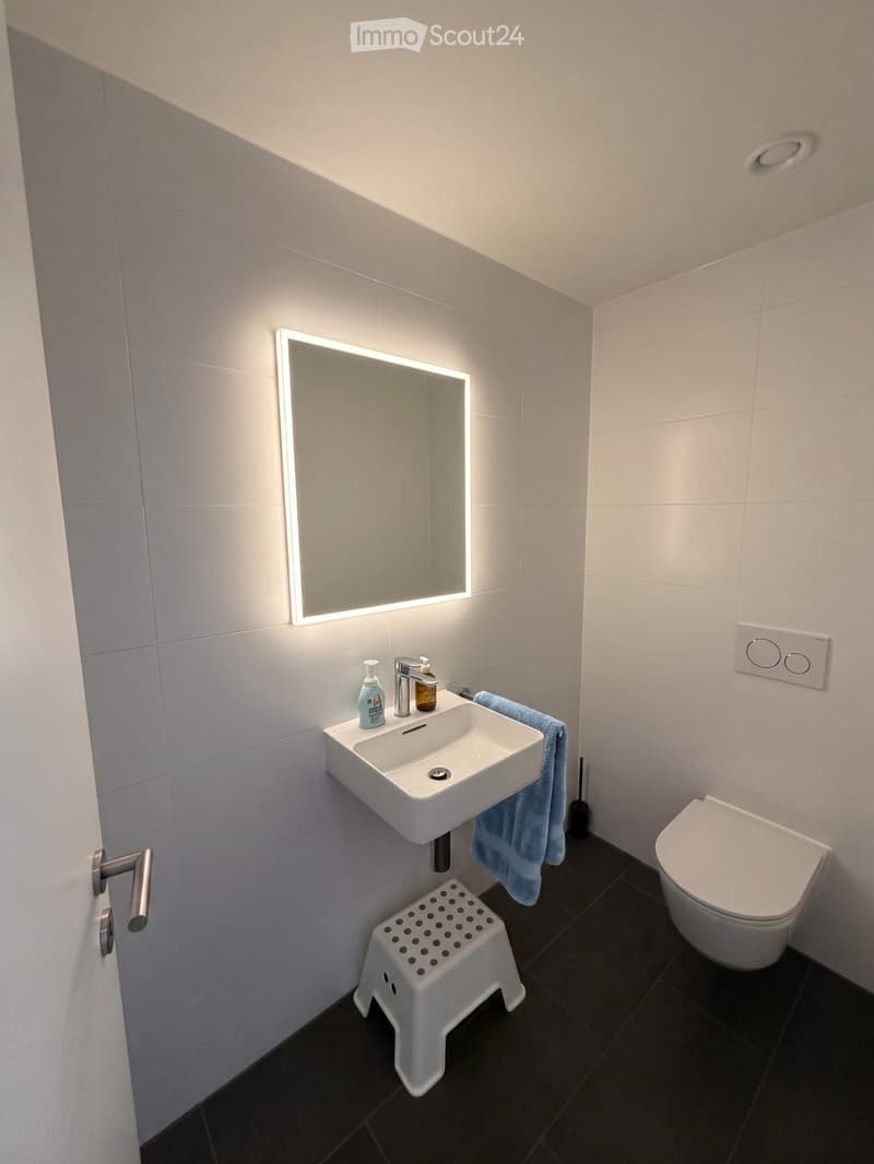 Modern bathroom with white tiles, a white sink, a mirror with LED lighting, a toilet, a small white stool, and a blue towel hanging on the side.