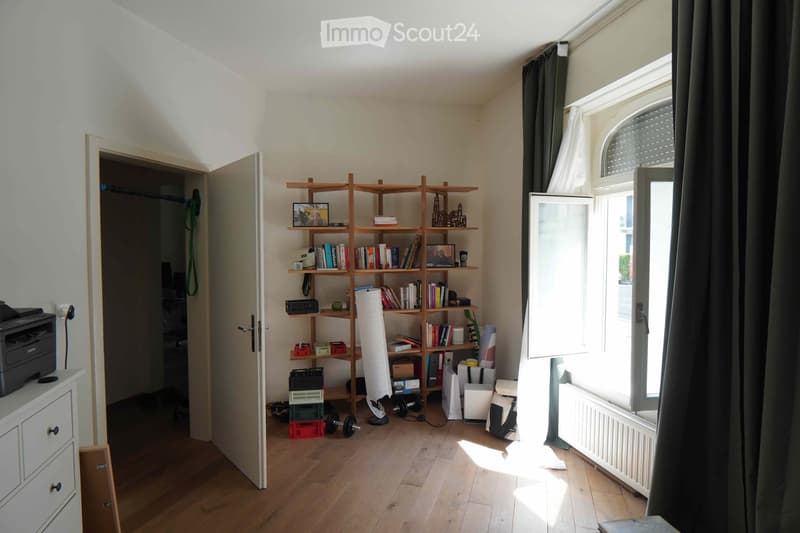 A well-lit room with wooden flooring, a large open shelving unit filled with books and decorative items, a printer on a small cabinet, and gym equipment near the window.
