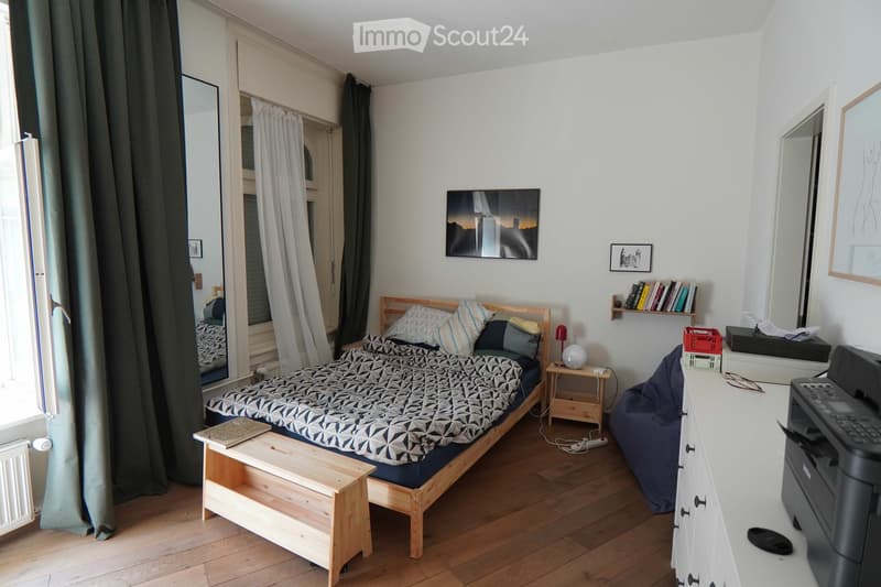 Cozy bedroom with wooden flooring, a single bed with patterned bedding, a nightstand with a lamp, a small wooden bench, a large mirror, a shelf with books, and a printer on a white desk.