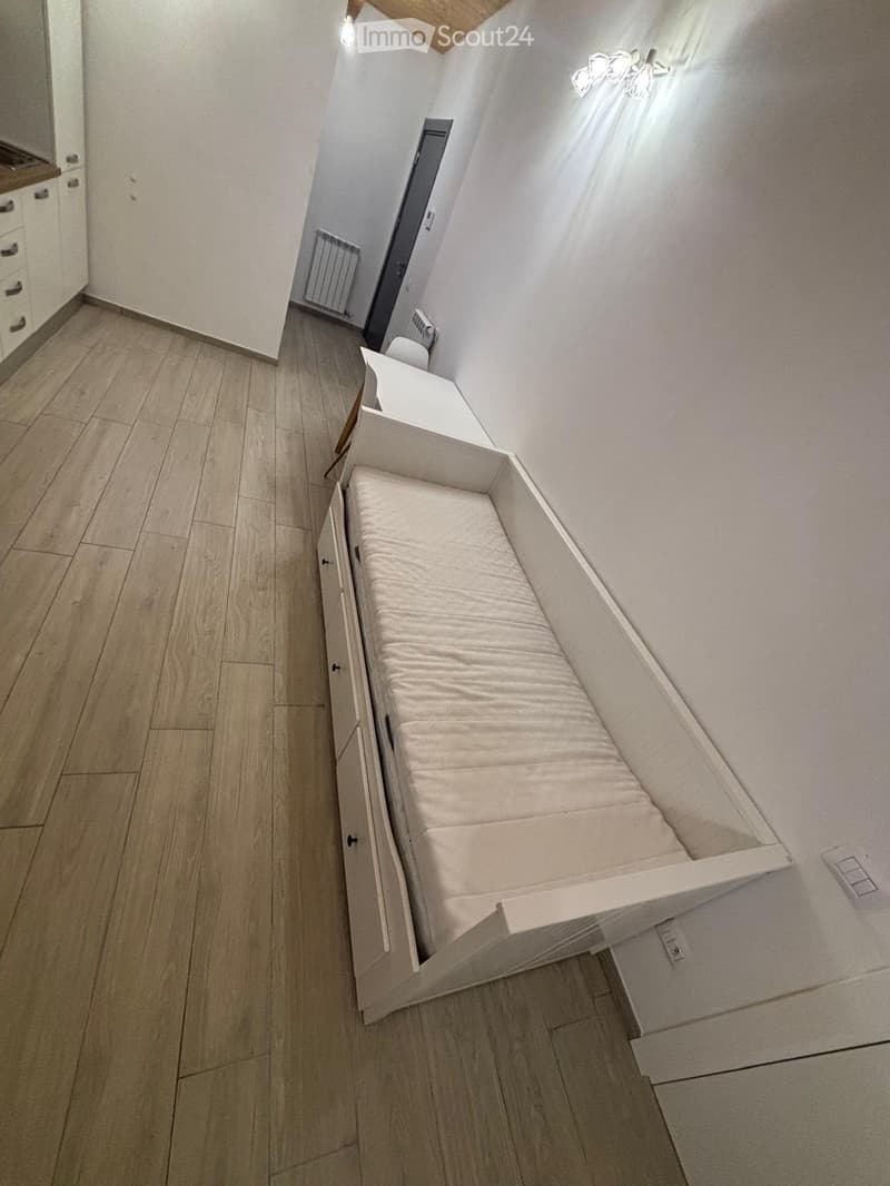 A modern, compact bedroom with a built-in sofa bed and a small cabinet, featuring light wooden flooring and white walls.