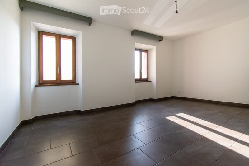 Empty room with dark tiled floor, white walls, and two windows with wooden frames. Natural light streams in from the windows, casting shadows on the floor.