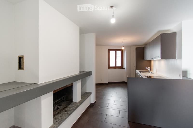 An unfurnished room with a fireplace, tiled floor, and a window. The space includes a kitchen area with cabinets and a sink, but no furniture or appliances are present.