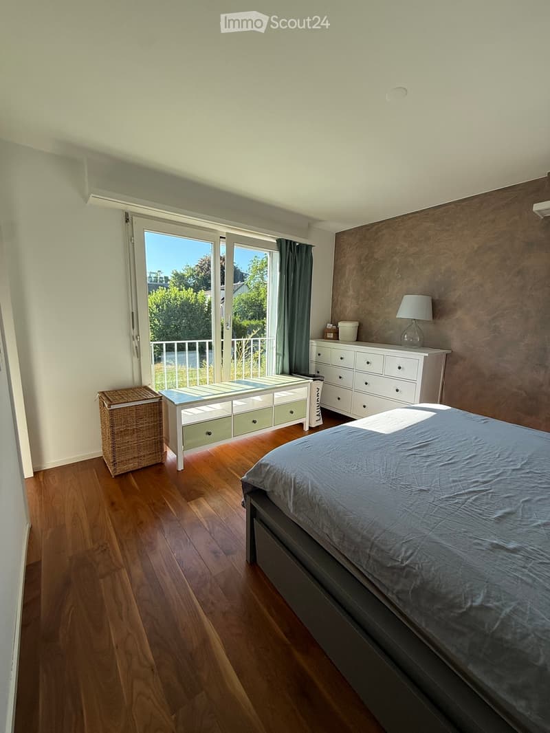 Cozy bedroom with wooden flooring, a large bed with grey bedding, a nightstand with a lamp, a drawer unit, a bench with storage, and a wicker basket near the sliding glass door leading to a balcony.