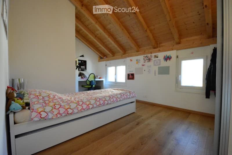 Bright bedroom with wooden floors and slanted wooden ceiling. Features a single bed with colorful heart-patterned bedding, a small desk with a chair, and a mirror on the wall.