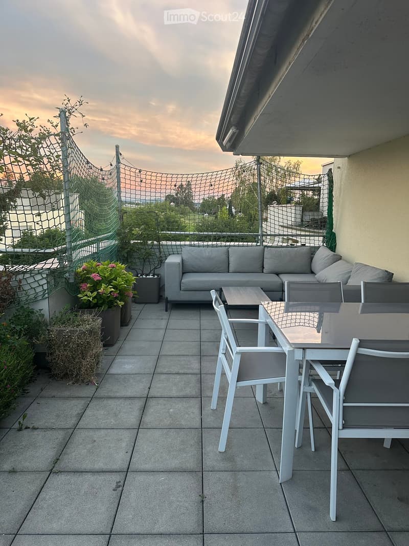 Balcony with grey sofa, small table, and white chairs, surrounded by potted plants and a net fence.