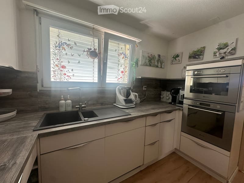 Modern kitchen with light-colored cabinets, a double sink, and stainless steel appliances including an oven and coffee maker. The countertops are made of wood, and there are decorative wall pieces above the cabinets.