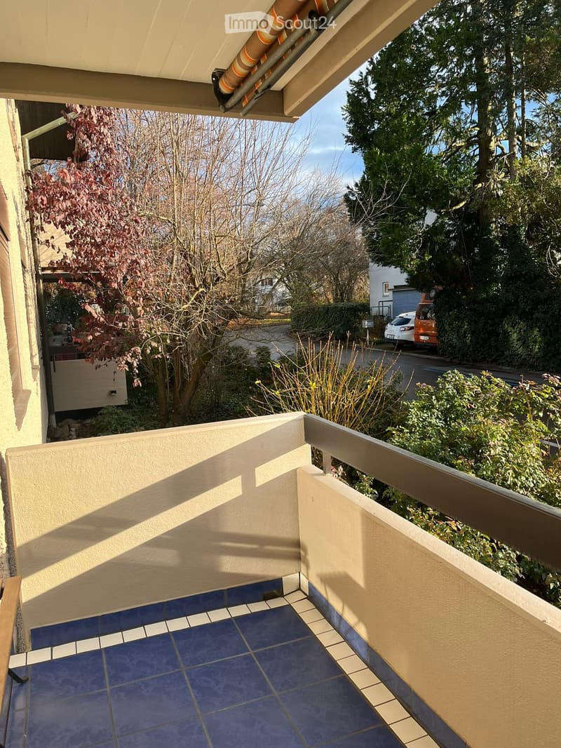 Balcony with blue tiled floor, beige walls, wooden railing, and view of trees and street.