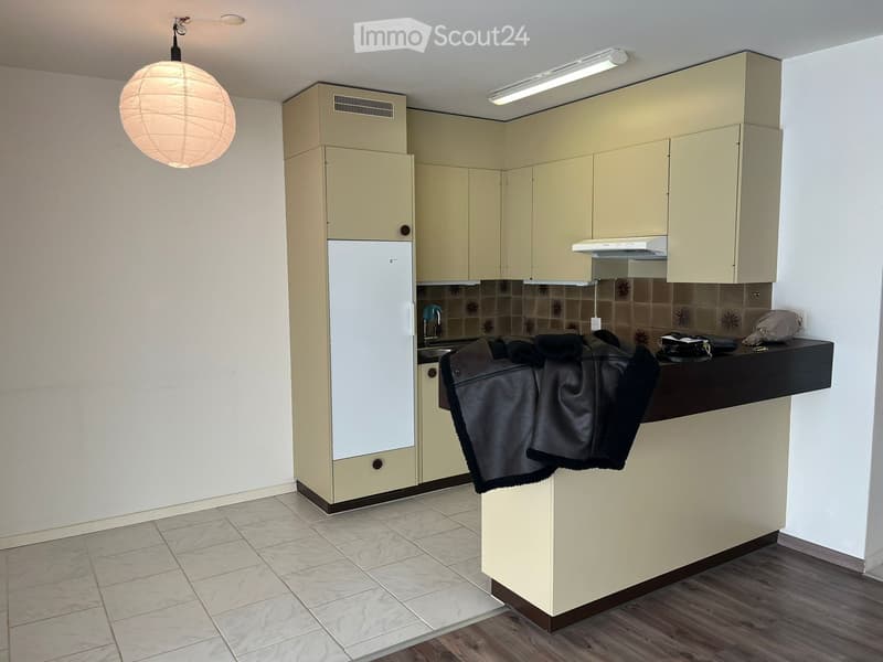Modern kitchen with light-colored cabinets, a refrigerator, a sink, and a range hood. A black countertop holds a folded jacket and some items. The floor is tiled, and the walls are painted white.
