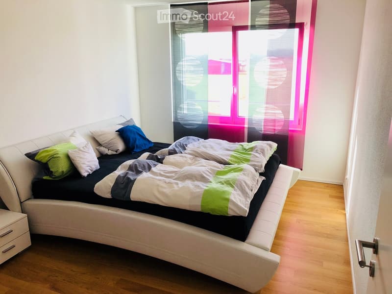 Modern bedroom with a white sofa bed, colorful pillows, and a blanket, positioned against a large window with sheer curtains.