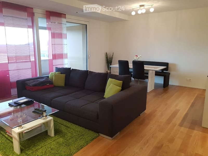 Modern living room with a dark grey sectional sofa, glass coffee table, dining area with a white table and black chairs, and a bench seating area.