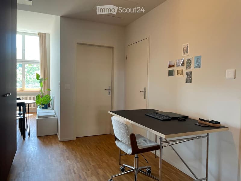 A modern, well-lit room with a wooden floor, a desk with a chair, a small table with stools, and a plant near the window. The walls are painted in neutral tones, and there are several framed pictures on one wall.