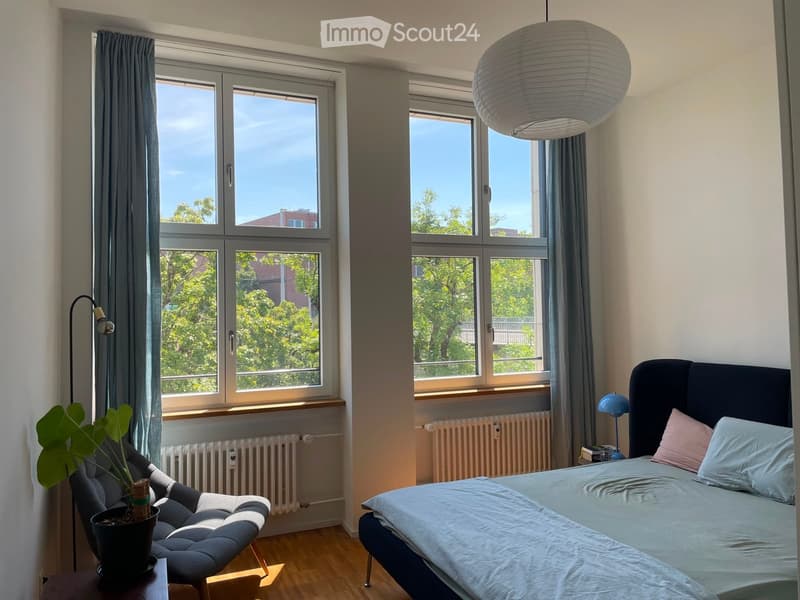 Bright bedroom with large windows, a double bed with light blue bedding, a grey armchair, a small table with a potted plant, and a hanging paper lantern light fixture.