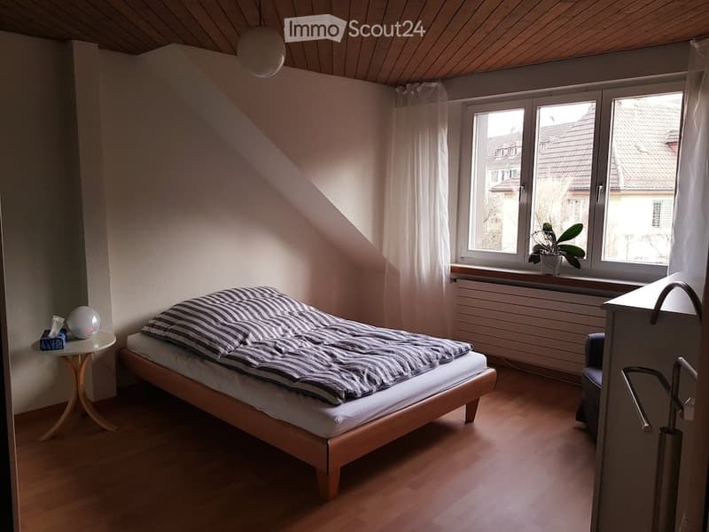 Cozy bedroom with wooden flooring, slanted ceiling, large window with white curtains, bed with striped bedding, small nightstand, and a plant on the windowsill.