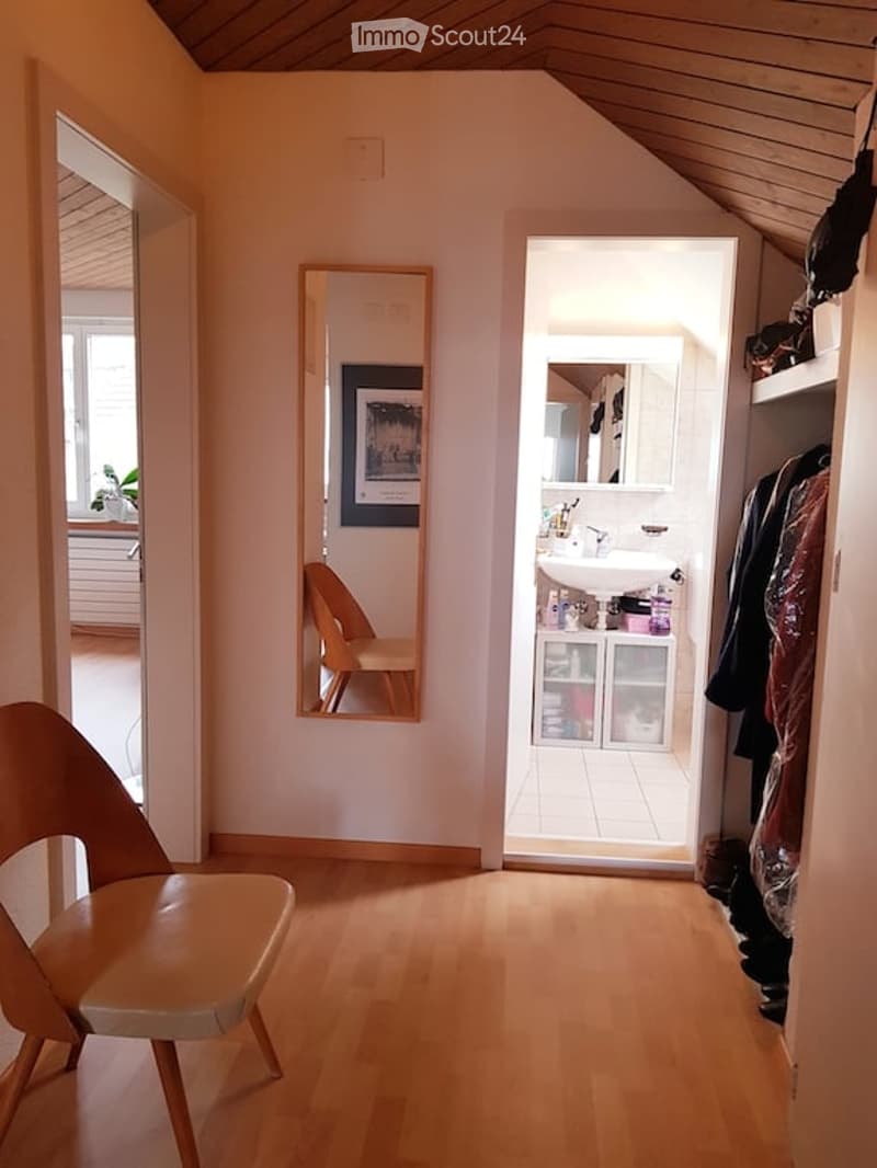 Bright, slanted-ceiling room with wooden flooring, a modern chair, a mirror, and an adjacent small bathroom with a sink and cabinet.