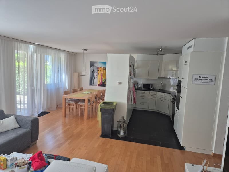 Open-plan living and dining area with a grey sofa, coffee table, dining table with chairs, modern kitchen with cabinets, refrigerator, microwave, and oven. Wooden flooring, large windows with curtains, and a painting on the wall.