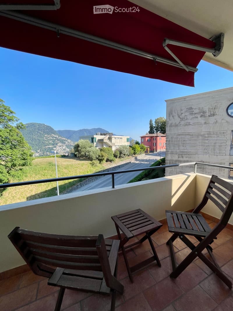 Balcony with wooden chairs and a small table, overlooking a scenic mountain view and town.