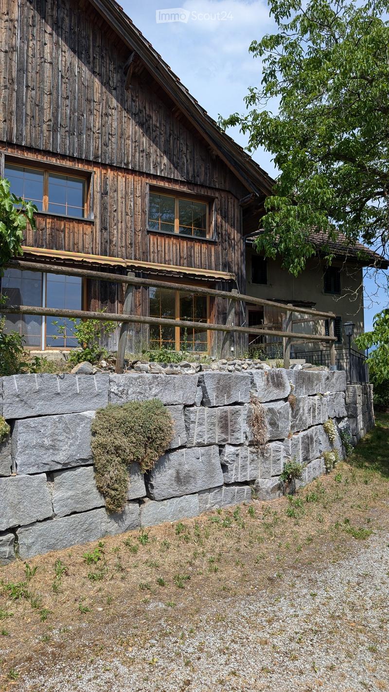 A rustic wooden house with a stone retaining wall in front, featuring large windows and a wooden balcony.