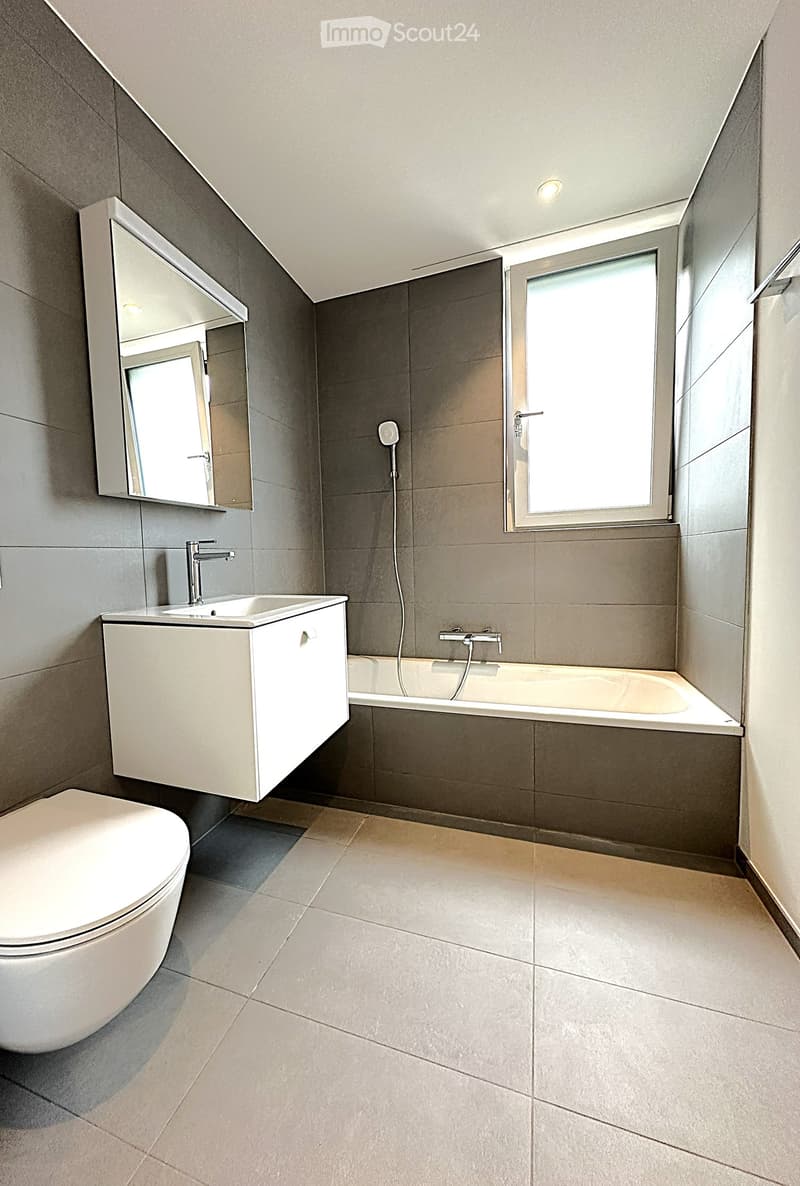 Bathroom. Modern bathroom with a toilet, sink, bathtub, mirror, and window. Grey tiles and white fixtures.