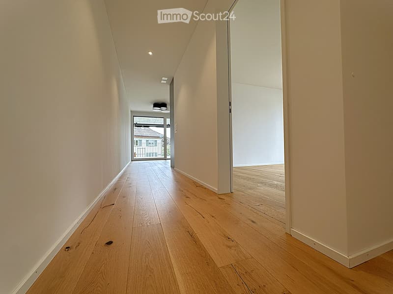 Corridor / Hallway. A narrow, well-lit hallway with light wooden flooring and white walls, leading to an open space with large windows and a view of the outside.