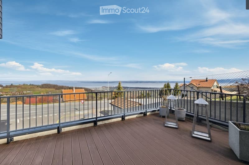 Balcony with wooden decking, metal railing, and potted plants, overlooking a scenic landscape with houses and a body of water.