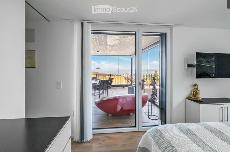 Modern bedroom with wooden flooring, large glass doors leading to a balcony with a scenic view, a bed, a red sofa, a dining table with chairs, and decorative statues.