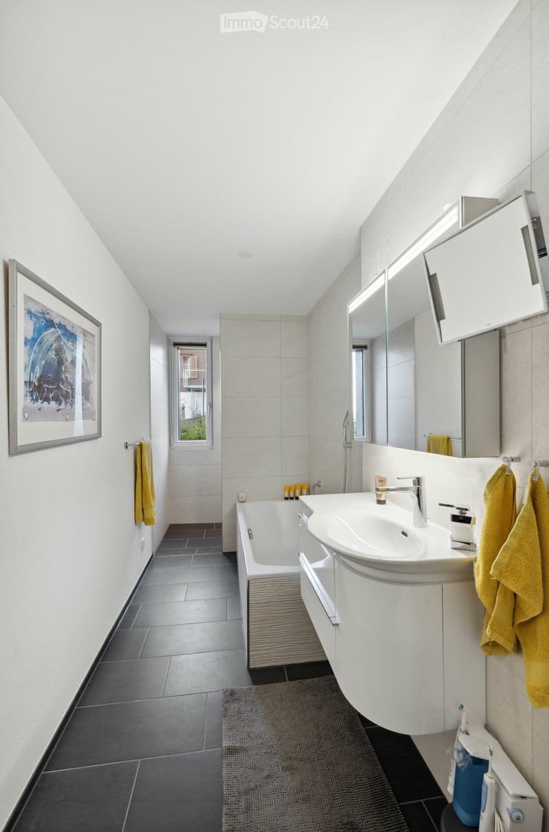 Modern bathroom with white tiles, a bathtub, a sink with a mirror, and a framed picture on the wall. Yellow towels are hanging on the sides.