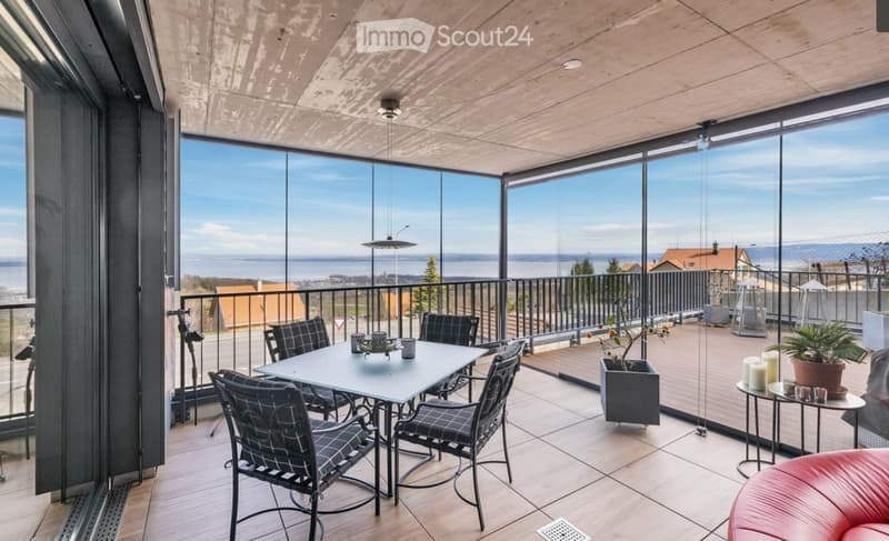 Modern balcony with large glass walls offering a scenic view, furnished with a dining table, chairs, potted plants, and a red sofa.