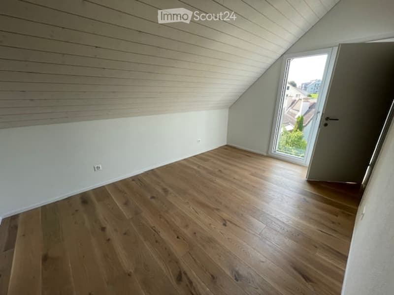 Empty attic room with wooden flooring, white walls, and a large window offering a view of the outside. The room has a slanted ceiling and a door leading to another area.