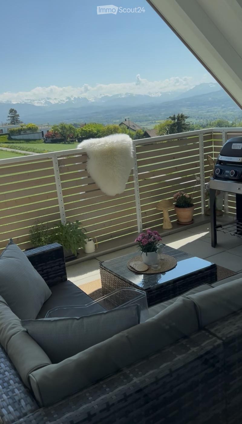 Balcony with a view of mountains, featuring a wicker sofa, a small table with flower pots, and a grill.