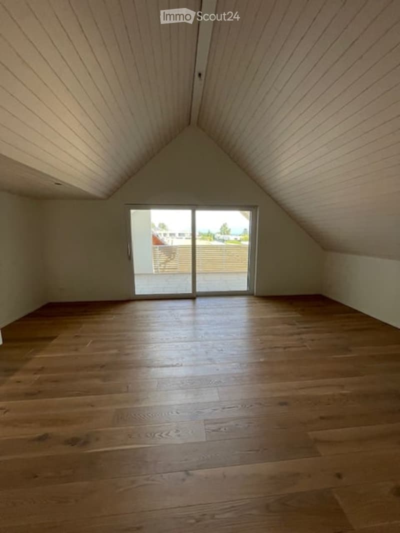 Bright, empty attic room with wooden floors, slanted wooden ceiling, and large sliding glass door leading to a balcony with outdoor view.