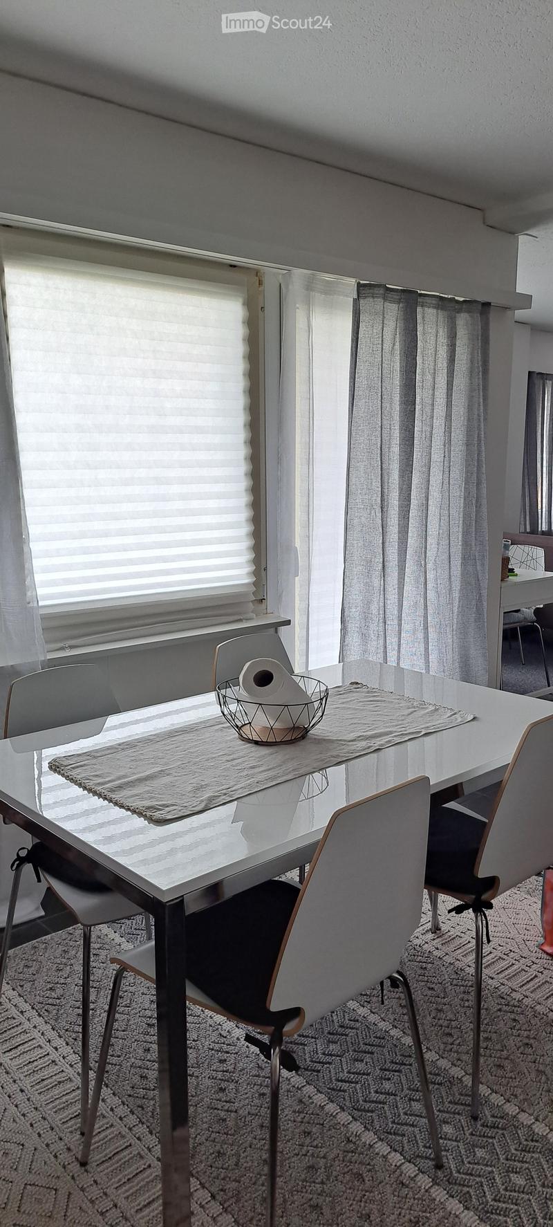 Dining area with a white table, four chairs, a woven placemat, and a basket with toilet paper. Large windows with blinds and curtains provide natural light.