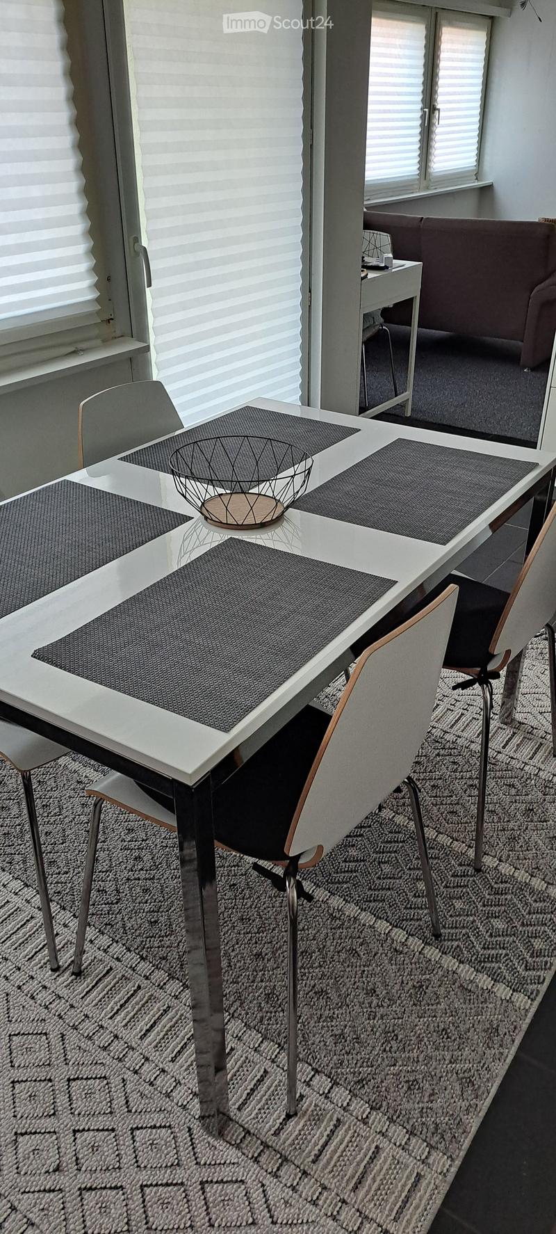 Modern dining area with a white dining table, four chairs, and a grey sofa in the background. The table is set with placemats and a decorative bowl.