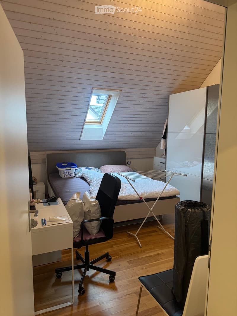 Cozy attic bedroom with wooden walls and slanted ceiling, featuring a bed with bedding, a desk with office chair, a laundry drying rack, and a small storage box on the bed.