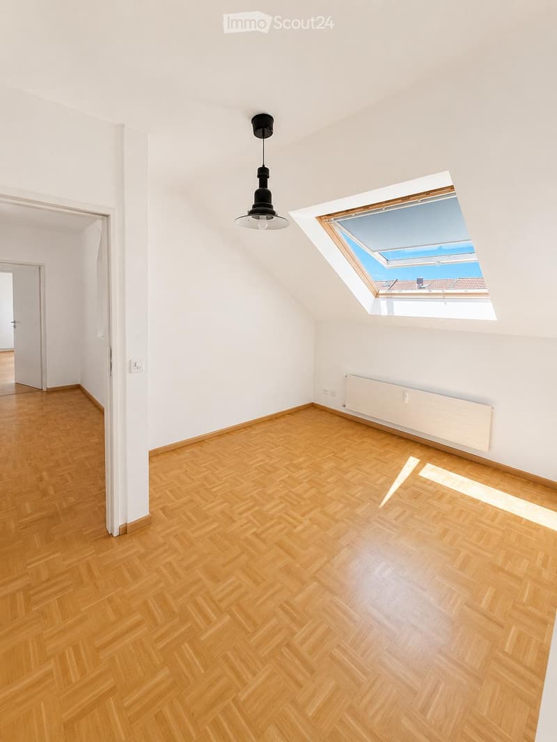 Empty room with wooden herringbone flooring, white walls, a skylight window, and a single hanging light fixture.