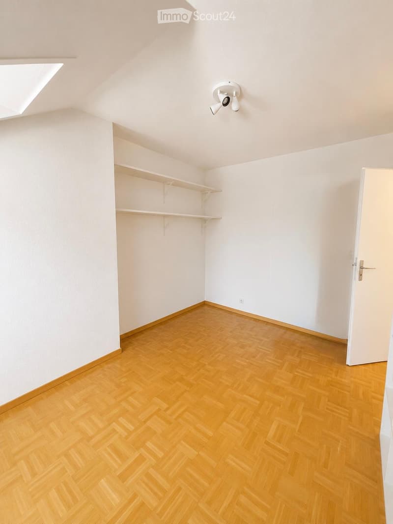 Empty room with wooden herringbone flooring, white walls, built-in closet with empty shelves, and a skylight window. No furniture or equipment present.