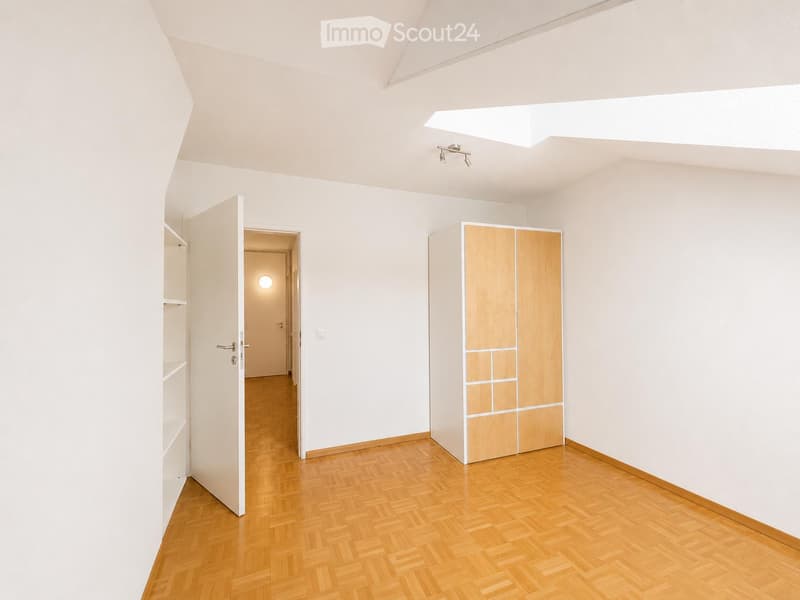 Empty room with light wooden herringbone flooring, white walls, and a built-in wooden closet. The room features a skylight and an open doorway leading to another space.