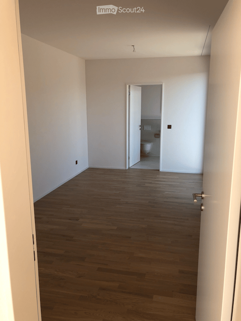Empty room with wooden flooring and white walls, featuring an open doorway leading to a small bathroom with a toilet.