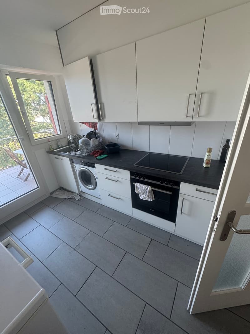 Modern kitchen with white cabinets, black countertop, induction stove, oven, washing machine, and sink. Features a large window with a view of greenery.
