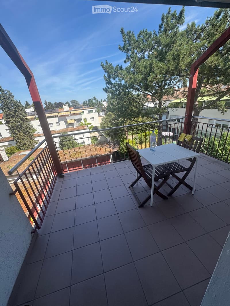 Balcony with tiled floor, metal railing, white table, wooden chairs, and a bottle on the table.