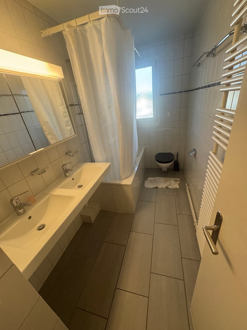 Modern bathroom with a sink, toilet, shower area, and a radiator. The space features tiled walls and floor, a large mirror above the sink, and a window providing natural light.