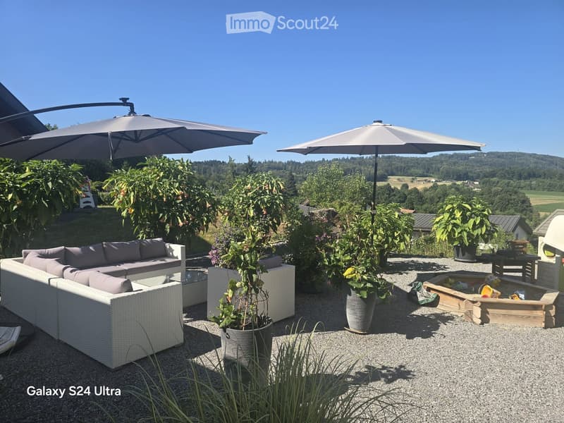 Outdoor patio with a white wicker sofa set, a wooden coffee table, two large umbrellas, and several potted plants. The area is surrounded by greenery and offers a scenic view of a valley.