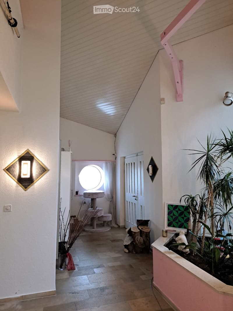 Bright hallway with tiled floor, white walls, decorative lighting fixtures, a small cat area with a round window, and a potted plant display on the right side.