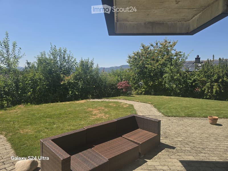 A well-maintained garden with a paved patio area, featuring a wicker sofa and table, surrounded by lush greenery and a clear blue sky.