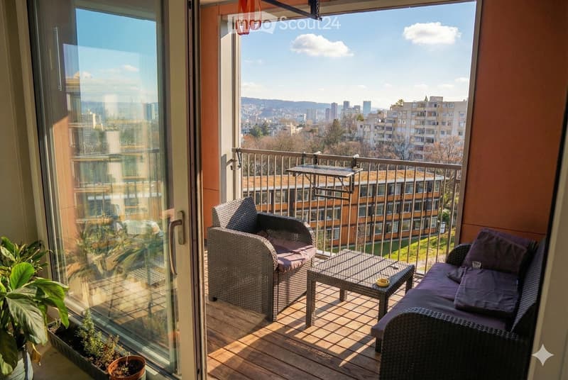Balcony with wooden decking, wicker furniture including a sofa, chair, and small table, and potted plants. Offers a scenic city view.